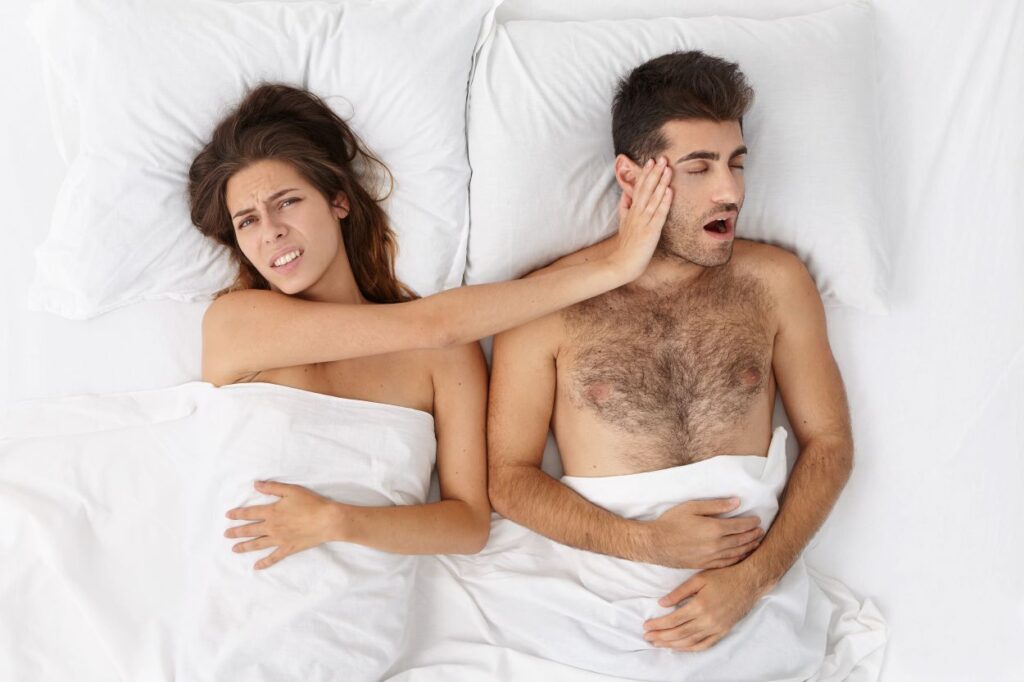 woman pushing away snoring partner in bed; snoring is a sleep apnea symptom