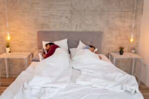 Couple lying together in the same bed back-to-back under separate duvets sleeping Scandinavian style.