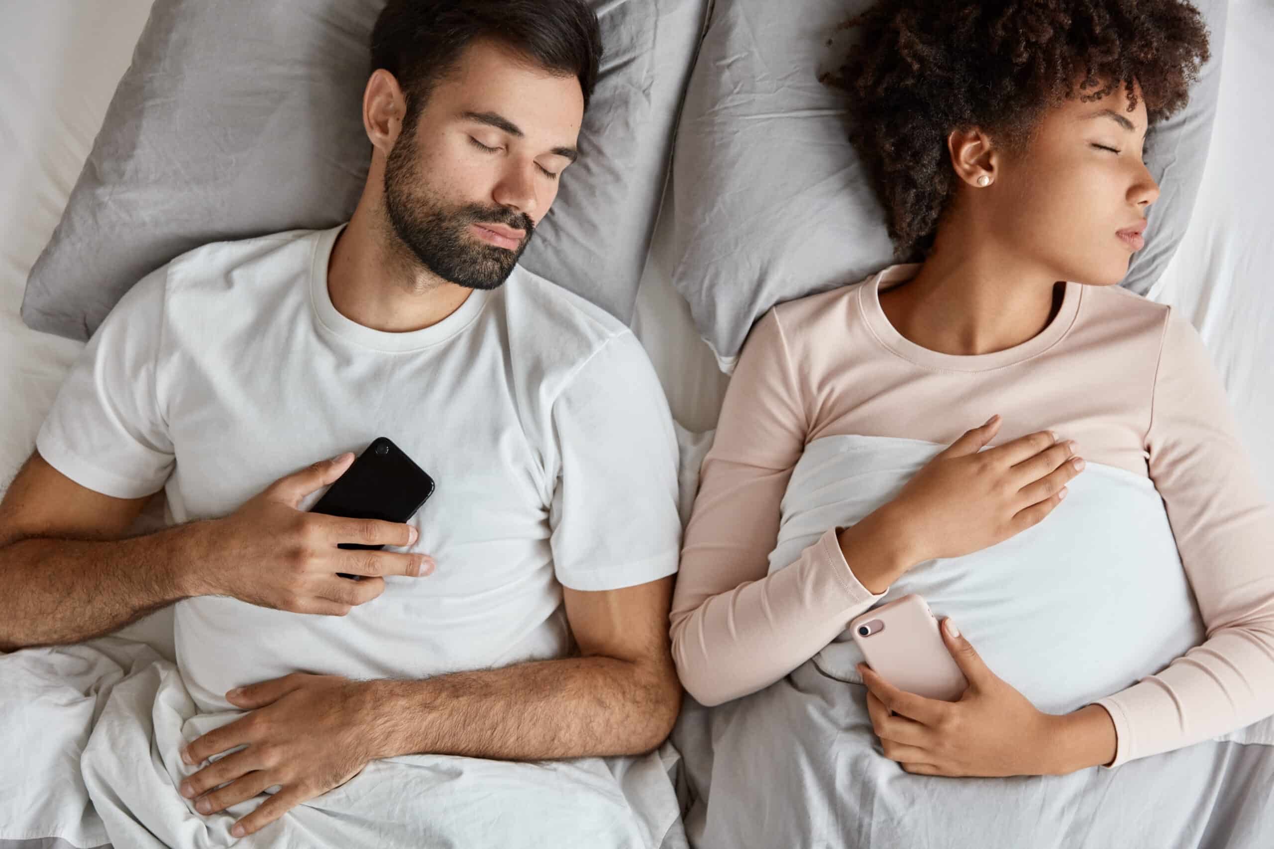 Top view of tech-addicted mixed race couple sleeping with their cell phones, modern electronic gadgets in hand.