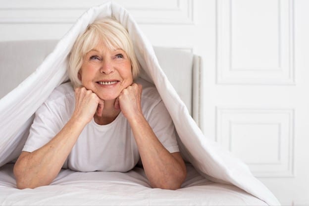 smiling senior woman in organic mattress with covers over top of head
